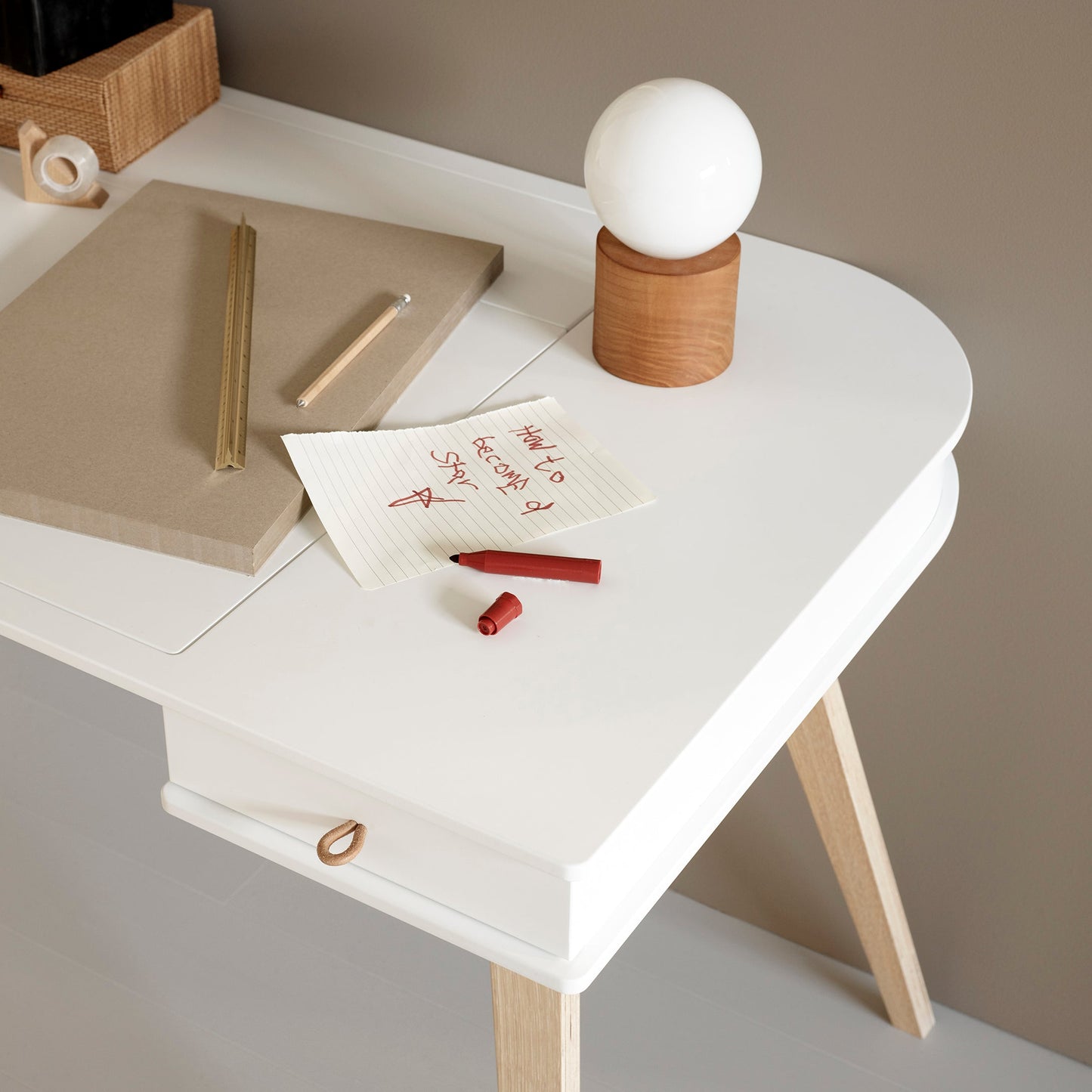 White desk with wooden legs, notebook, pen, and small lamp on a neutral background