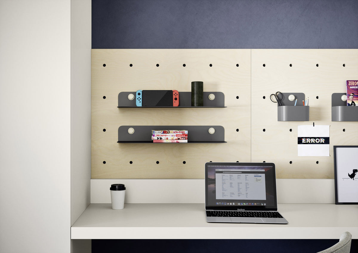 A modern office teen desk setup featuring a light wood pegboard wall with black floating shelves, gray wall hooks, and a white desk surface. A laptop, coffee cup, and framed print are arranged on the desk against a dark navy and white wall background.