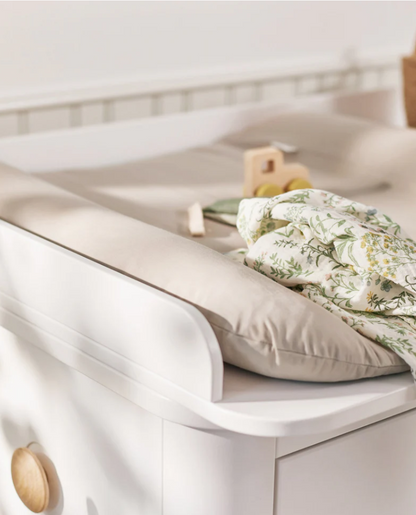 White crib with beige pillow and green floral blanket in a softly lit room
