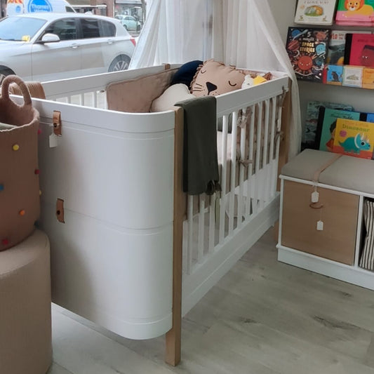 A white wooden baby crib with a white canopy netting, styled in a nursery room with a window showing a street view, toy shelves, and a hot air balloon decoration.
