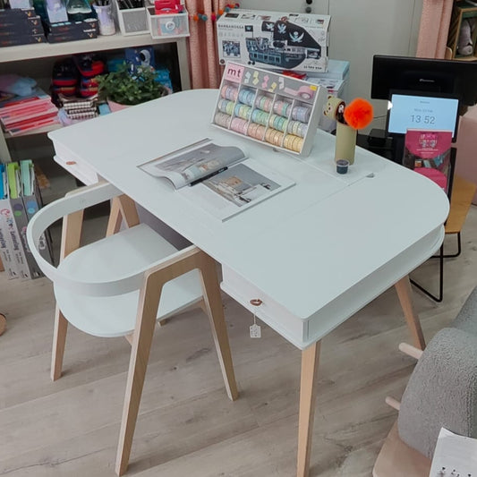A white rectangular writing desk with light wood legs, accompanied by two white chairs with light wood legs, set in a child's playroom with toys and storage shelves in the background.