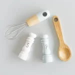 White salt shaker with wooden handle and whisk, and wooden spoon on white background