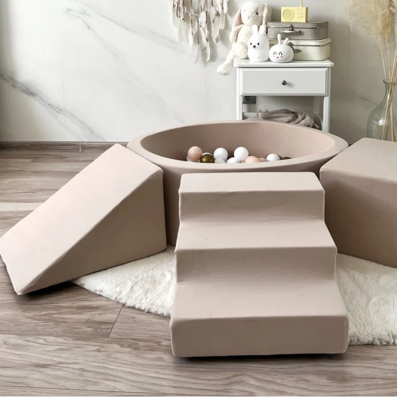 A soft play or pet climbing set made of beige fabric, including a ball pit with white and beige balls, a ramp, a staircase, and a cube, arranged on a white rug in a modern room with marble walls and wood flooring.