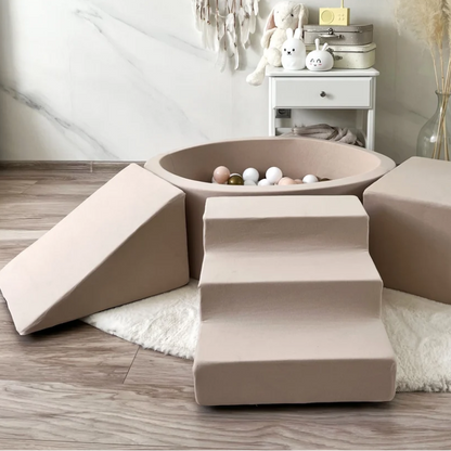 A soft play or pet climbing set made of beige fabric, including a ball pit with white and beige balls, a ramp, a staircase, and a cube, arranged on a white rug in a modern room with marble walls and wood flooring.