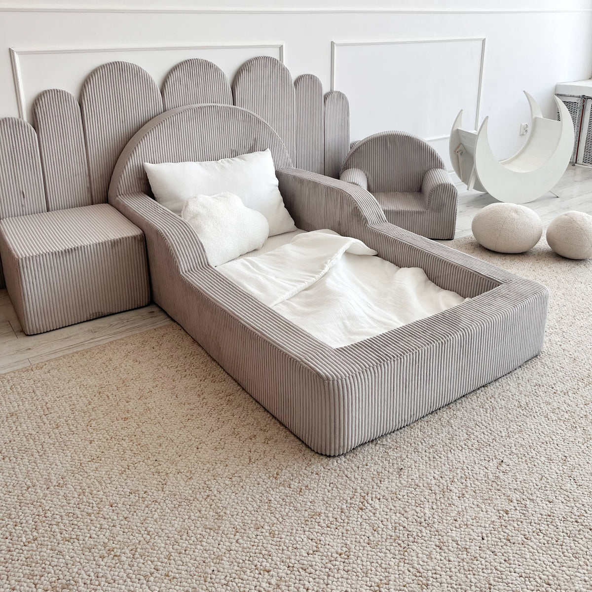 A modern children's bed in a light grey ribbed fabric, styled with white bedding and pillows, set against a white paneled wall in a bright room.