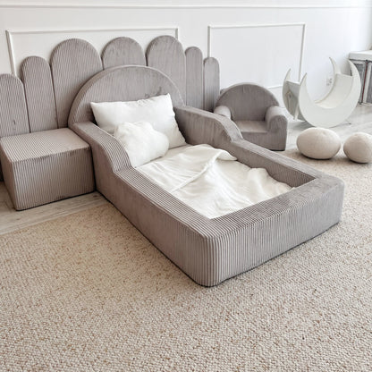 A modern children's bed in a light grey ribbed fabric, styled with white bedding and pillows, set against a white paneled wall in a bright room.