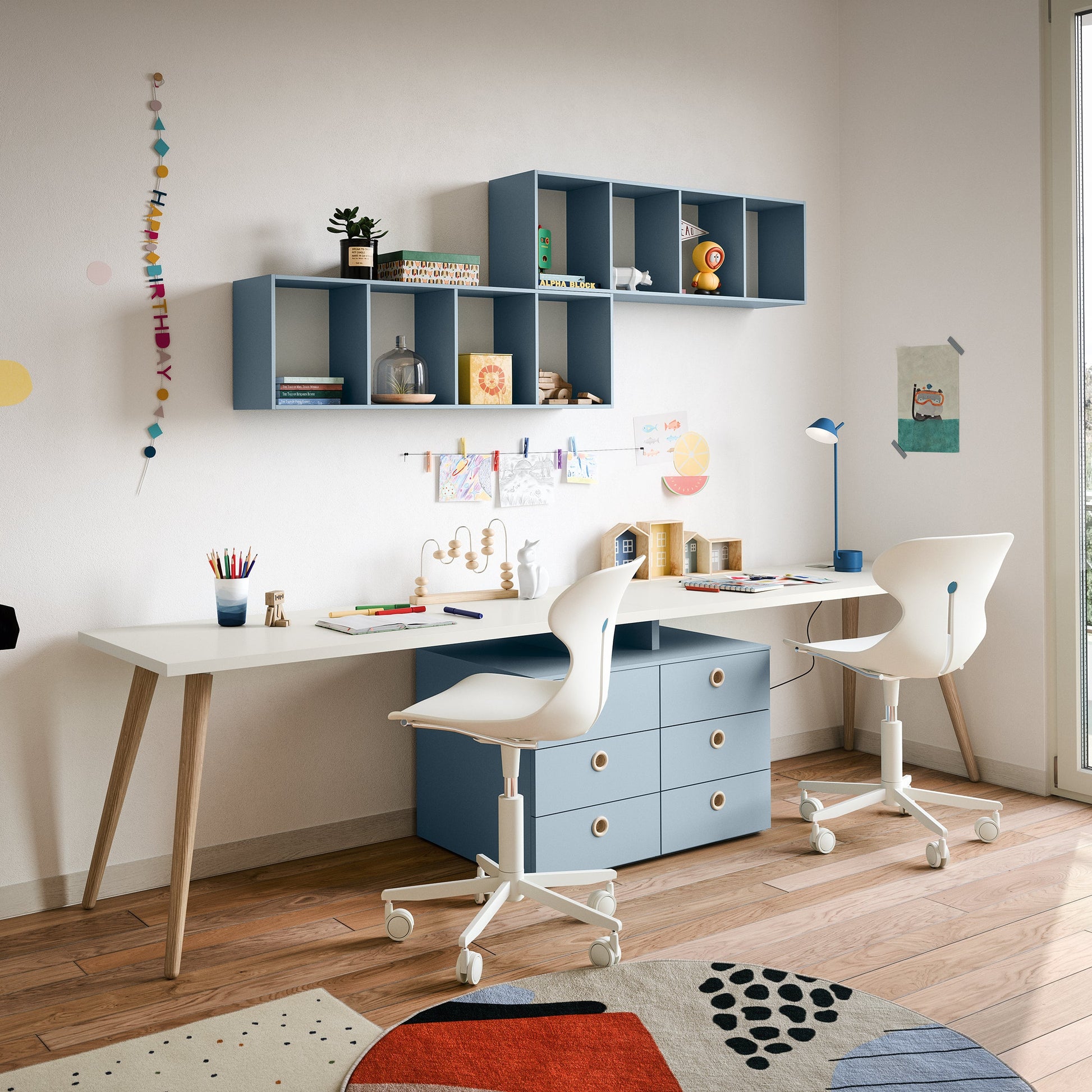 Shared kids study area with long desk, modular wall shelves, and blue drawer units in a Nidi room.