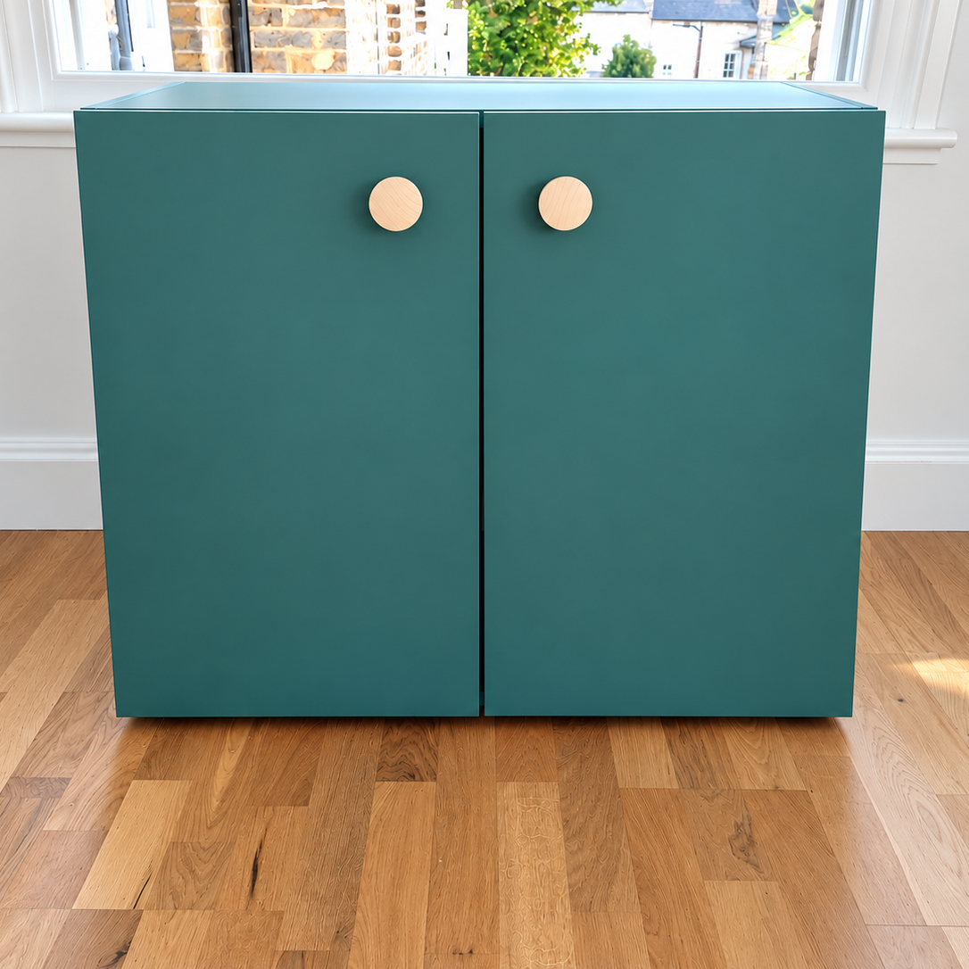A modern two-door cabinet in a matte teal green finish with round, light wood knobs, set against a white wall with a window and light oak flooring.