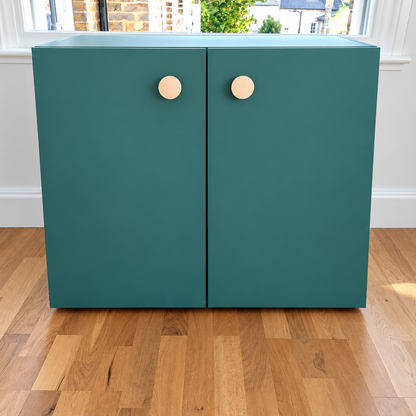 A modern two-door cabinet in a matte teal green finish with round, light wood knobs, set against a white wall with a window and light oak flooring.