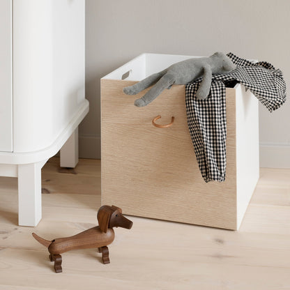 Wooden laundry basket with clothes on a wooden floor next to a white cabinet.