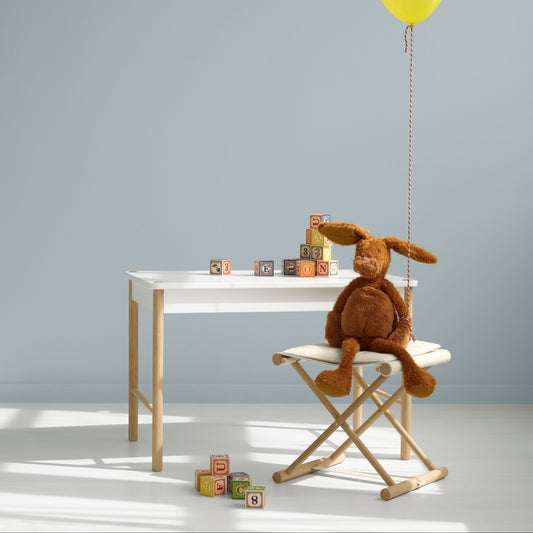 A brown plush bunny toy sits on a white folding chair next to a white table, with a bright yellow balloon floating above it against a pale blue-gray wall.