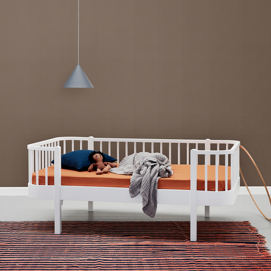 White junior daybed with wooden mattress against a brown wall with a pendant light.