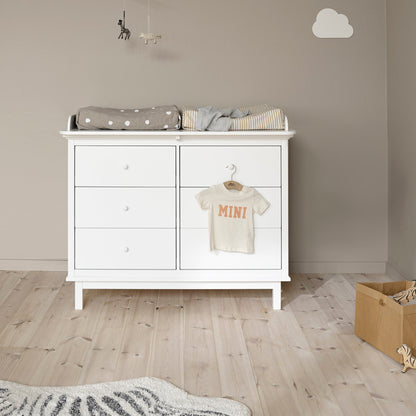 A white wooden changing table or dresser with three drawers on the left and two larger doors on the right, shown in a styled nursery. A folded gray polka-dot blanket and a striped cloth are placed on top, and a small cream baby T-shirt with orange text hangs from a hook on the right door.