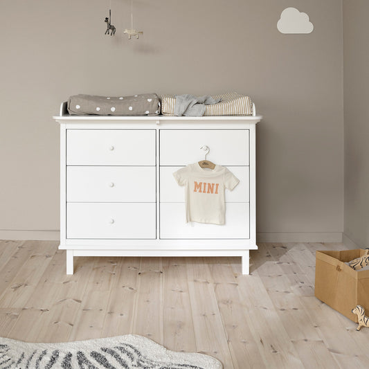 A white wooden changing table or dresser with three drawers on the left and two larger doors on the right, shown in a styled nursery. A folded gray polka-dot blanket and a striped cloth are placed on top, and a small cream baby T-shirt with orange text hangs from a hook on the right door.