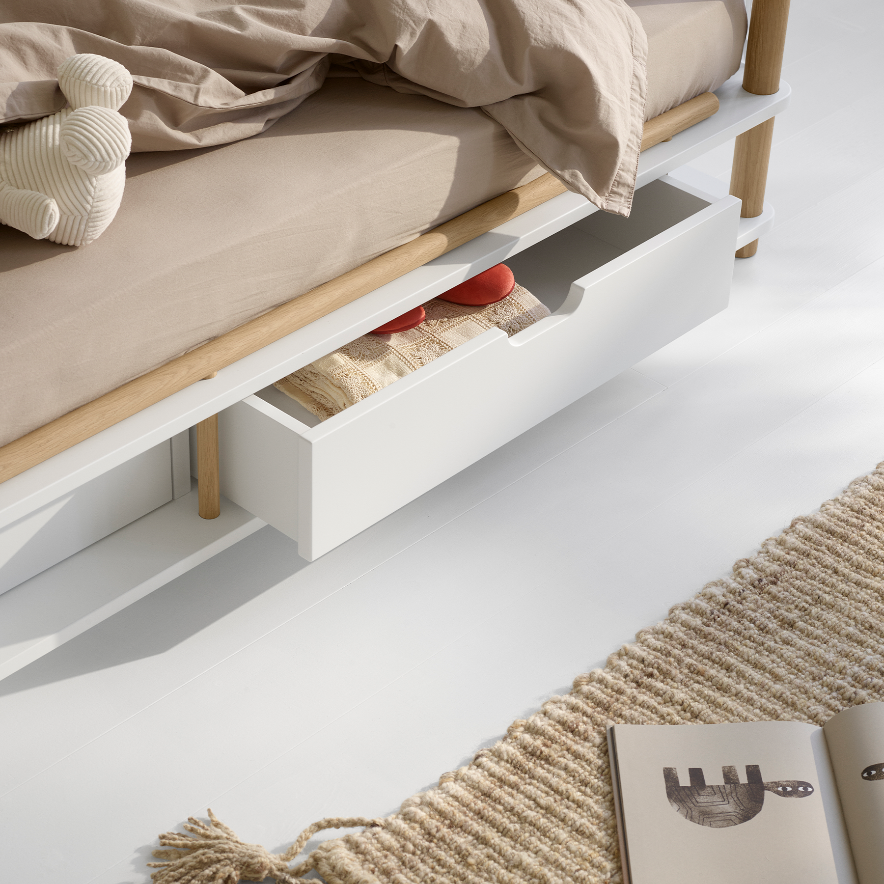 Wooden bed with beige bedding and a small drawer, on a white floor with a rug.