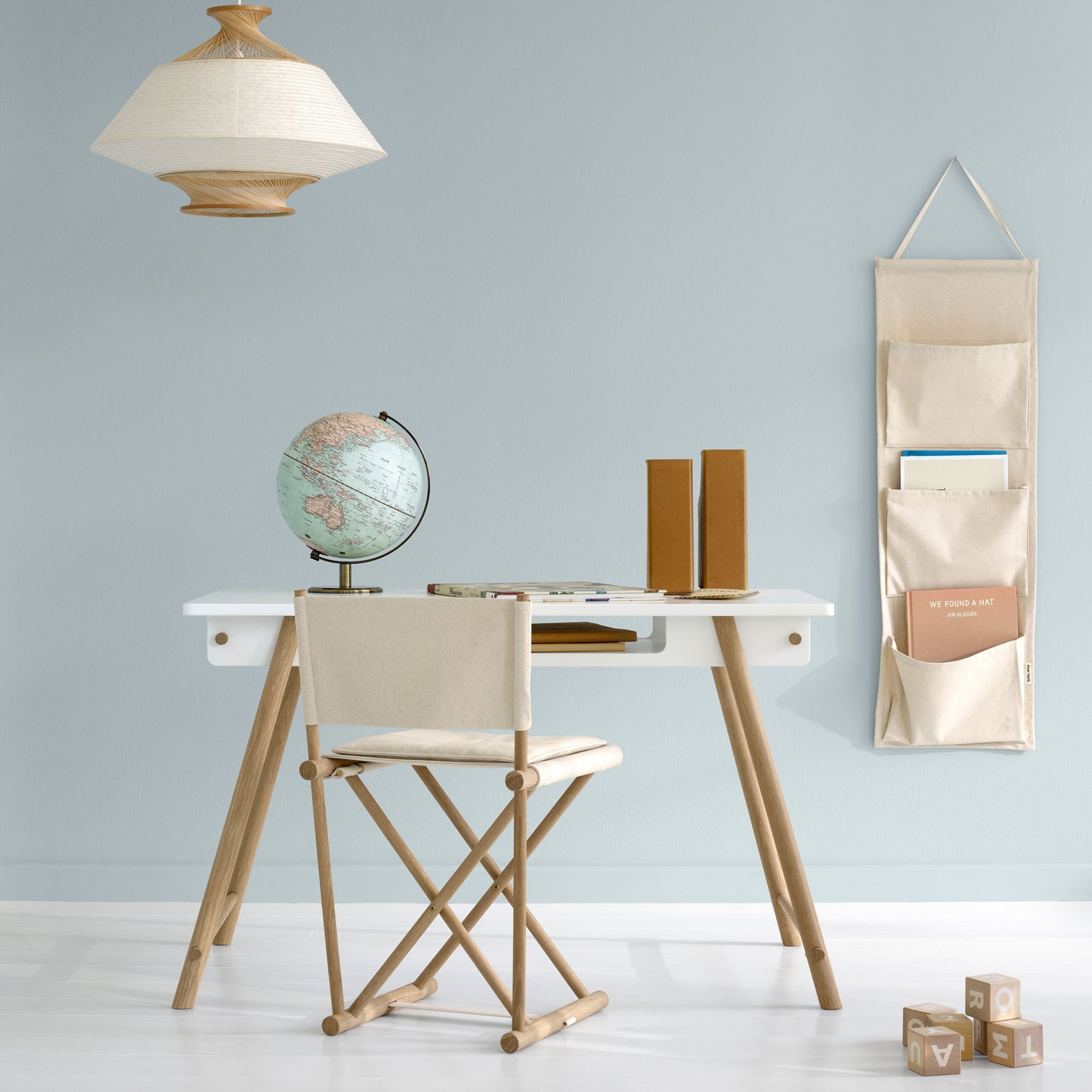 A styled interior scene with a white desk, light wood legs, a cream fabric chair, a hanging beige pocket organizer on the wall, a pendant lamp, and a globe on a pale blue wall.