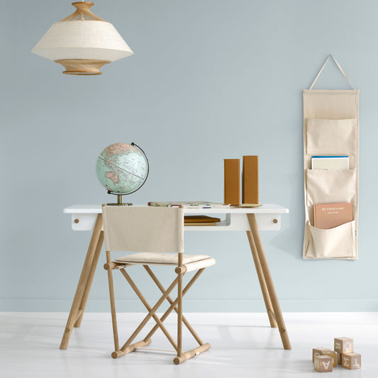A styled interior scene with a white desk, light wood legs, a cream fabric chair, a hanging beige pocket organizer on the wall, a pendant lamp, and a globe on a pale blue wall.
