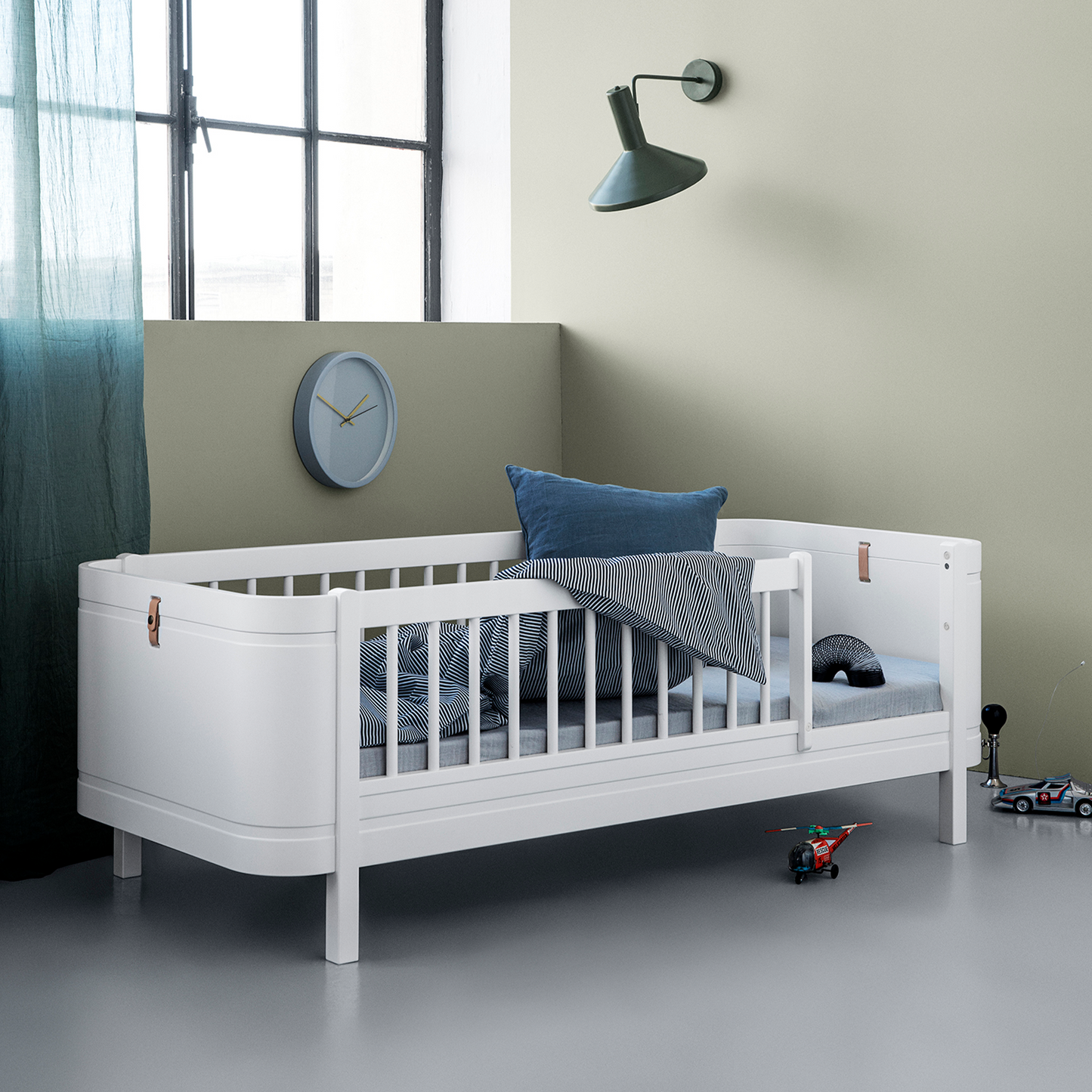 White crib with gray bedding in a room with a clock on the wall.