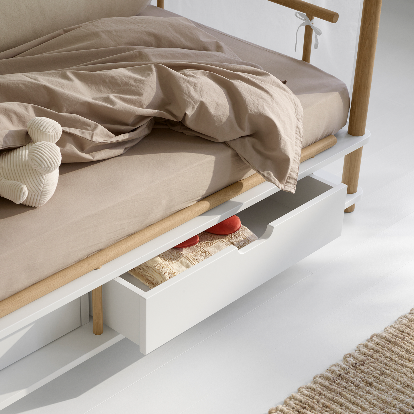 Wooden bed with beige bedding and a white drawer below on a light gray floor.