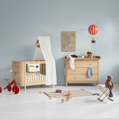 Oliver Furniture Nursery room with wooden crib, dresser, and toys against a light blue wall.