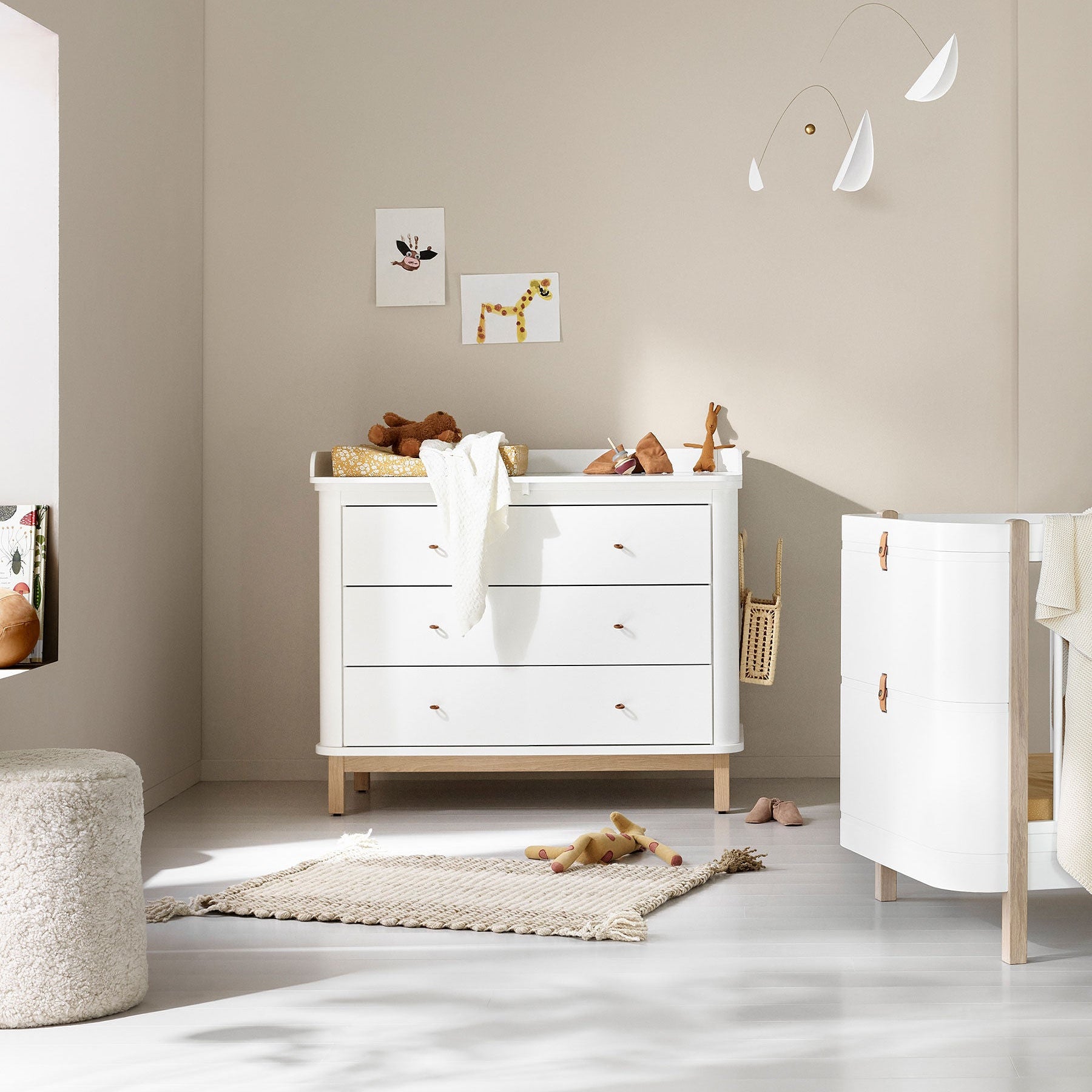 Nursery room with white dresser and crib, toys on the floor, and light-colored walls.