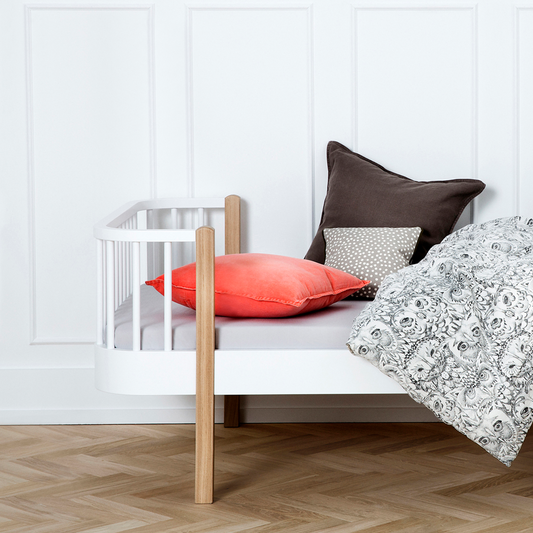White bed with wooden legs, adorned with colorful cushions and a patterned blanket against a white paneled wall.