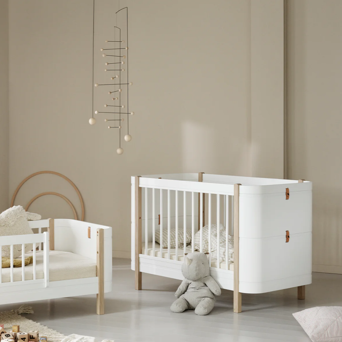 Children's room with white crib and wooden bed, beige walls, and toys on a light gray floor.