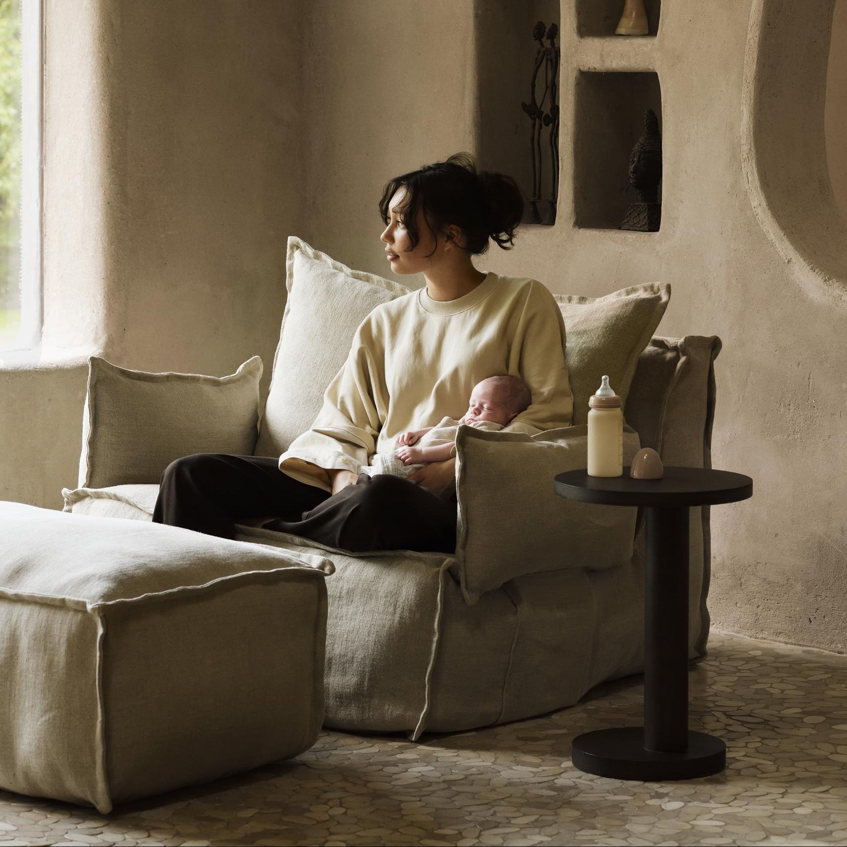 Woman sitting on a couch holding a baby in a cozy room with neutral tones.