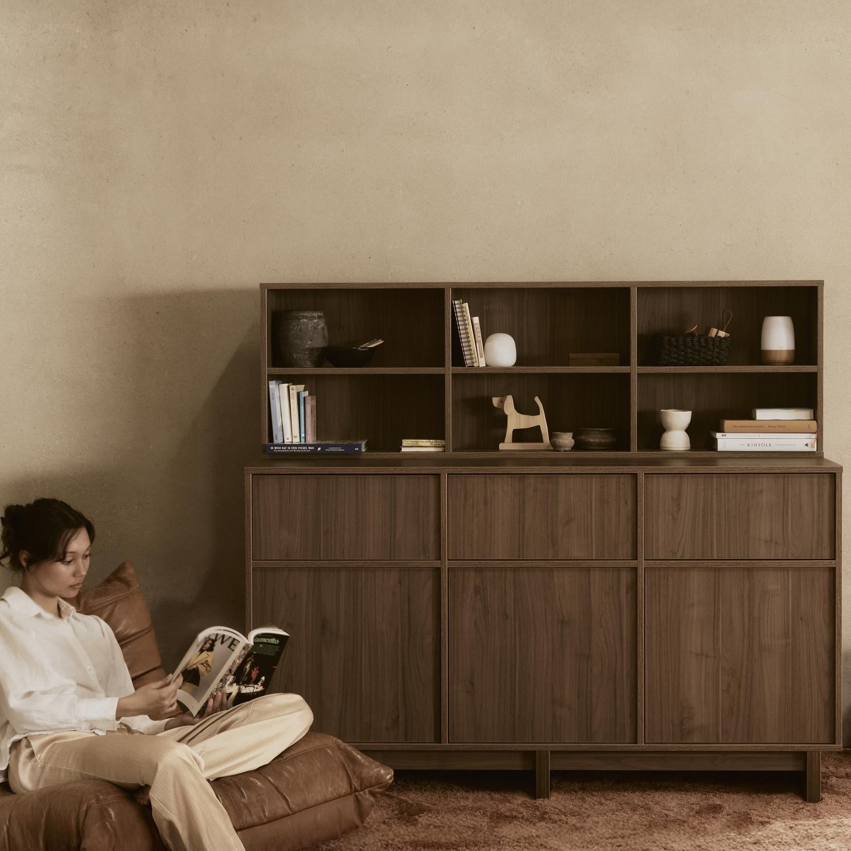 Woman sitting on a brown leather sofa reading a magazine in a room with a wooden cabinet.