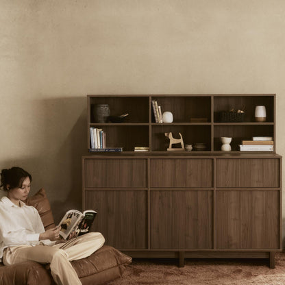 Woman sitting on a brown leather sofa reading a magazine in a room with a wooden cabinet.