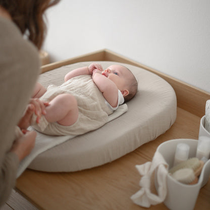 Baby lying on a white cushion with a woman beside them, in a room with a white wall and wooden floor.