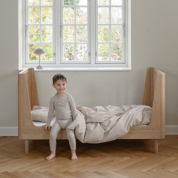Child sitting on a wooden bed in a room with large windows.