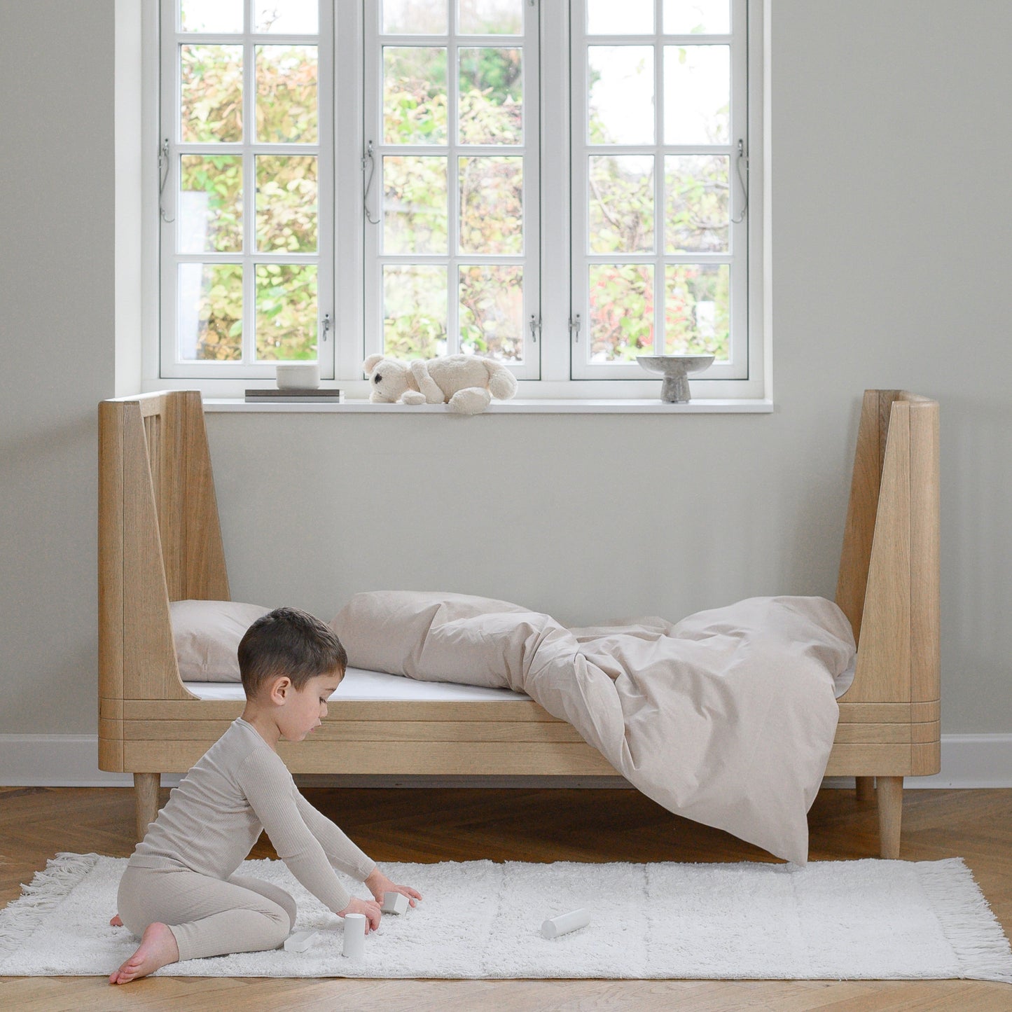 Child playing on a rug in front of a wooden crib with a window in the background