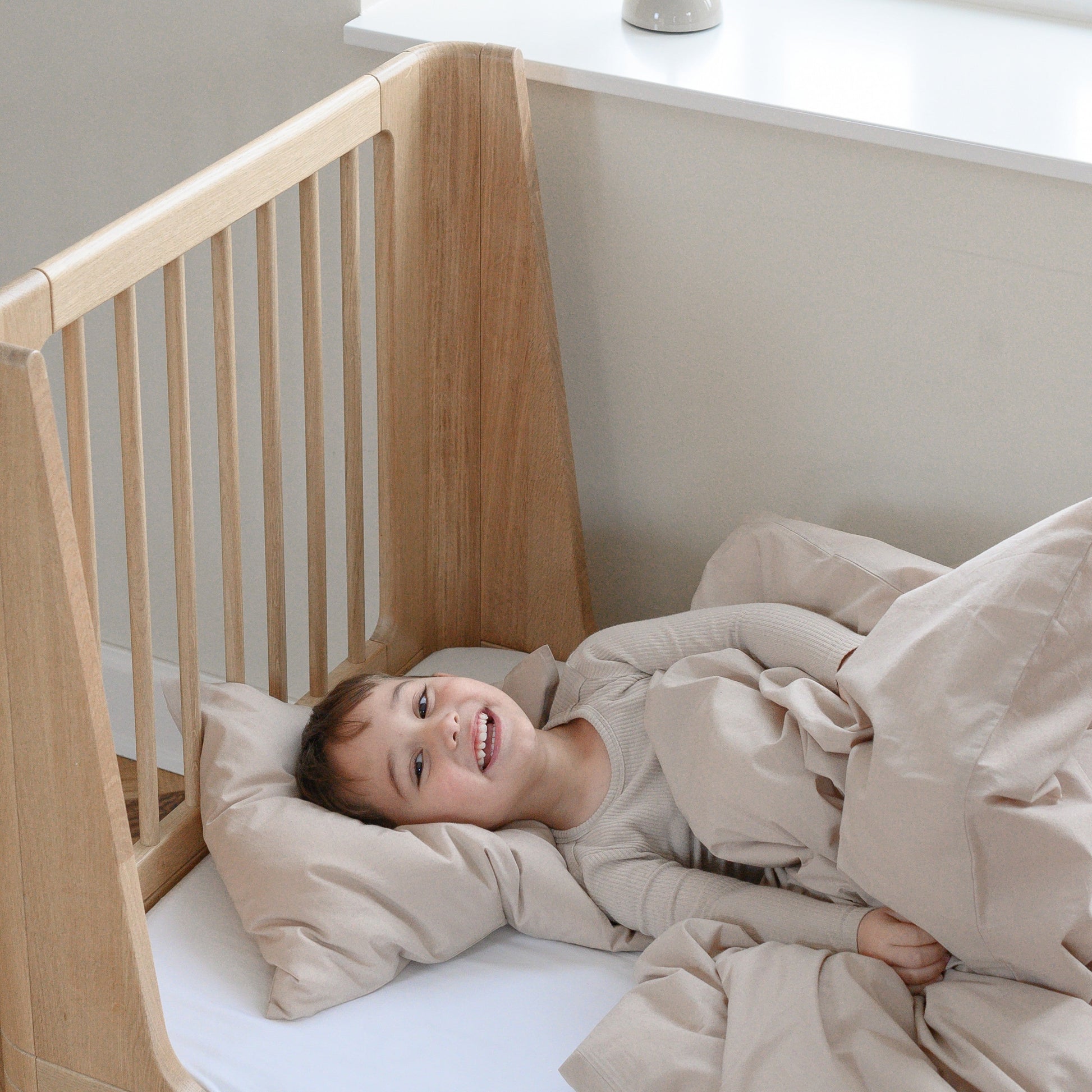 Child lying in a wooden oak crib with beige bedding, smiling.