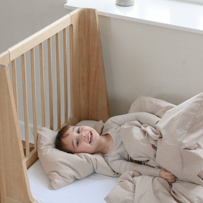 Child lying in a wooden oak crib with beige bedding, smiling.