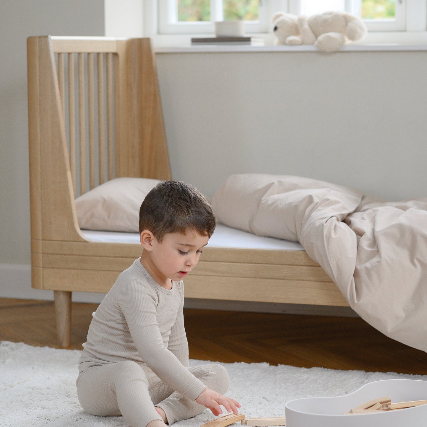 Child playing with toys on the floor in a bedroom with a crib and window in the background.
