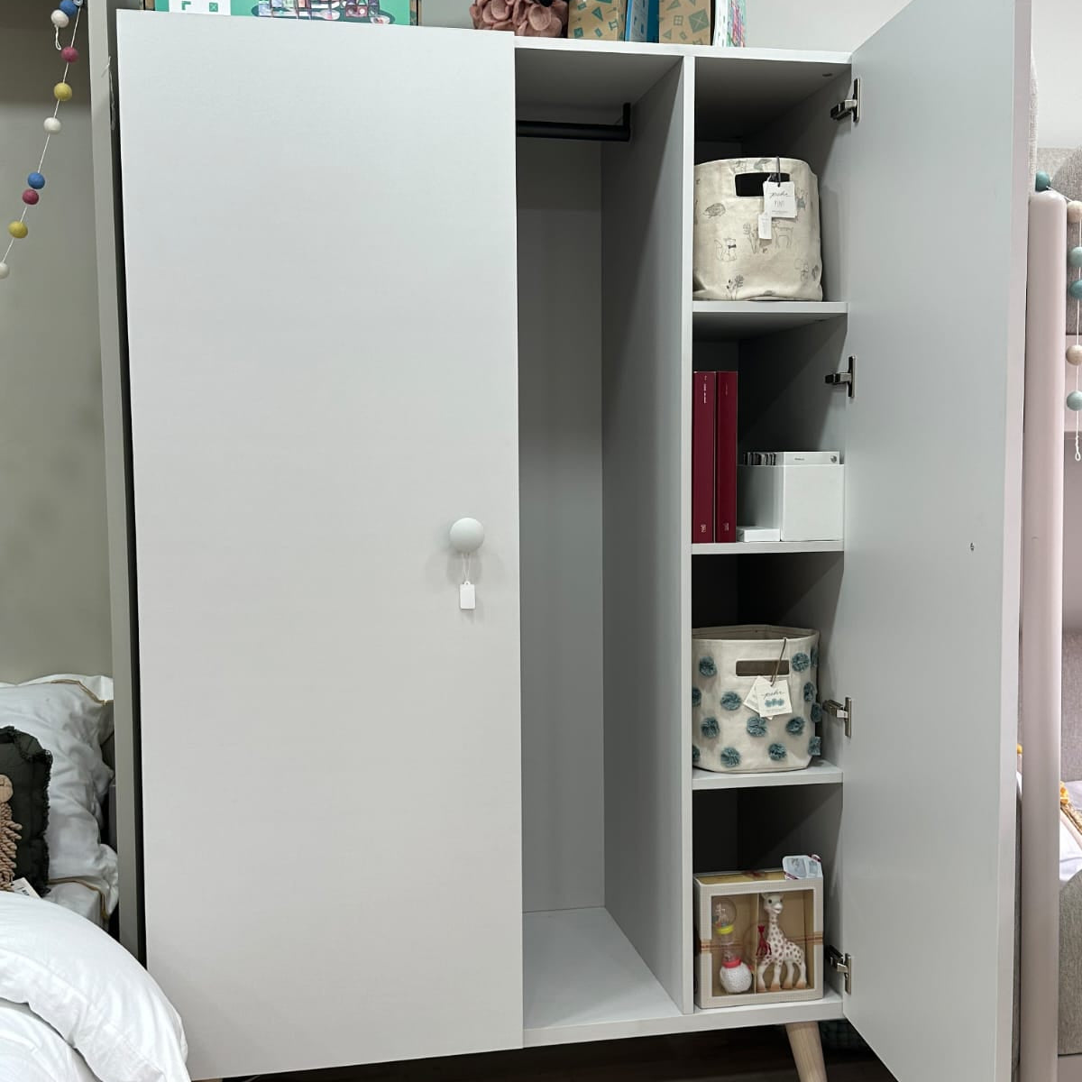 A tall white wardrobe or storage cabinet with two doors, one open to show shelves and storage bins inside, photographed in a bedroom setting.