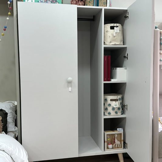 A tall white wardrobe or storage cabinet with two doors, one open to show shelves and storage bins inside, photographed in a bedroom setting.