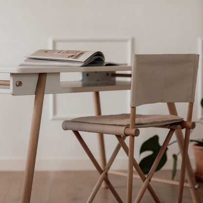 A minimalist light wood folding desk paired with a matching wooden director-style chair with beige fabric seat and back, set in a bright, modern room with a white wall and wood floor.