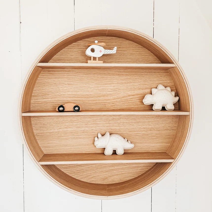 Round wooden nursery shelf with three tiers, styled with small wooden toy animals including dinosaurs and a helicopter, set against a white wood paneled wall.