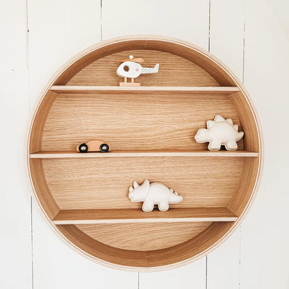 Round wooden nursery shelf with three tiers, styled with small wooden toy animals including dinosaurs and a helicopter, set against a white wood paneled wall.