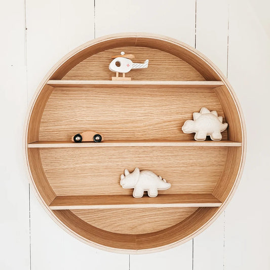 Round wooden nursery shelf with three tiers, styled with small wooden toy animals including dinosaurs and a helicopter, set against a white wood paneled wall.